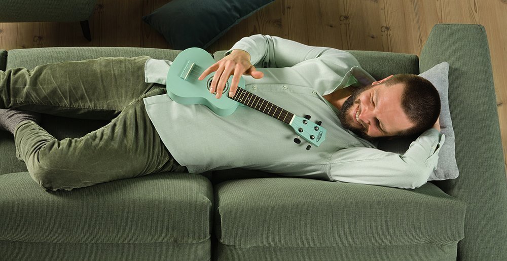 A young man lies on the sofa and has it good. He plays a small guitar and enjoys the moment.