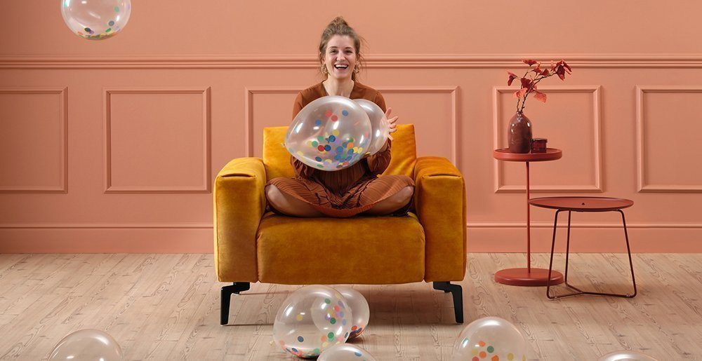 Een jonge vrouw zit met haar benen gekruist op een gele Cosy1 1-zitbank en speelt met ballonnen. Ze lacht. Hieruit moet blijken hoe gemakkelijk het is om het comfort van Sensoo aan te passen.